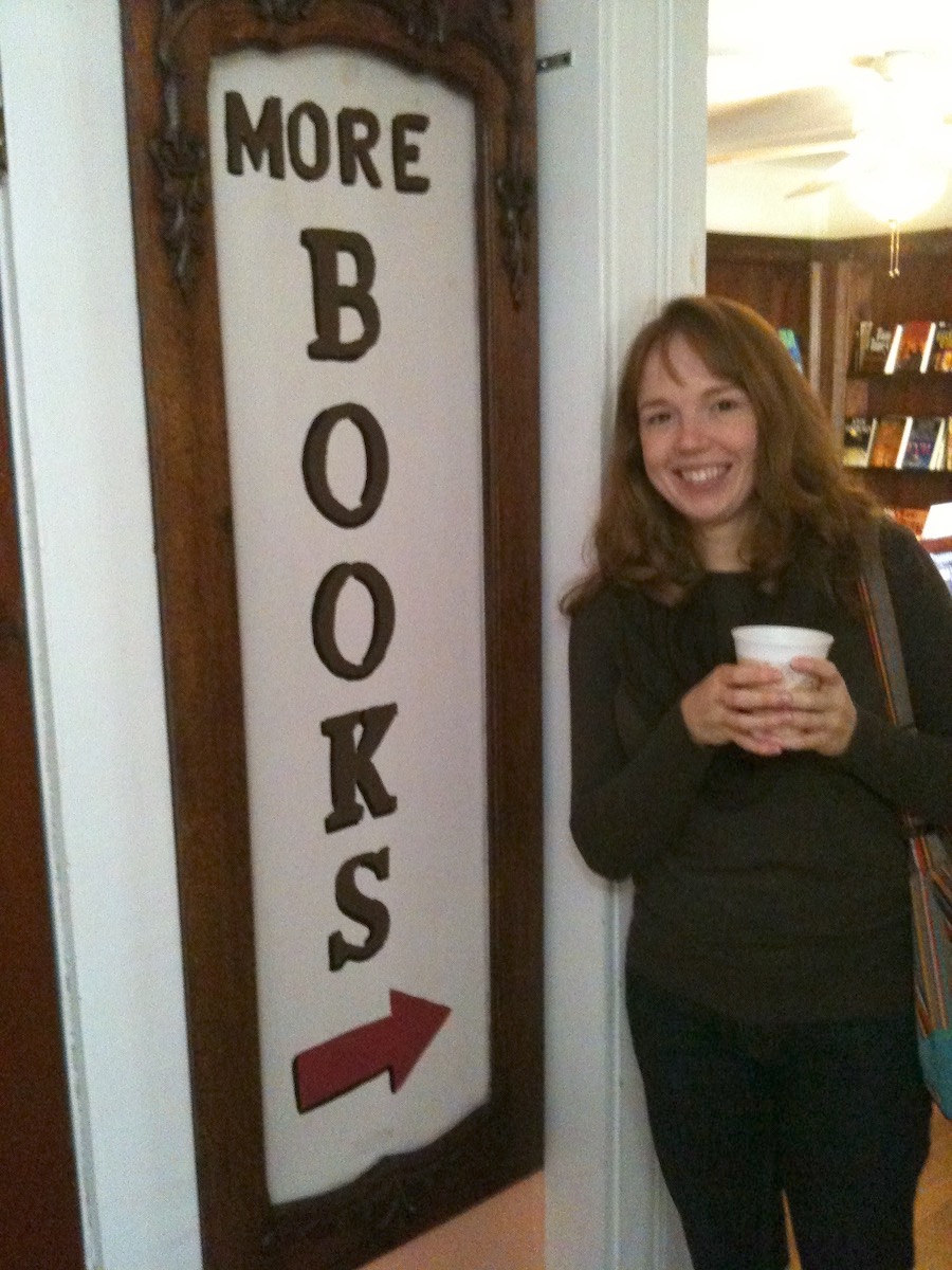 Amy holding a cup of coffee at a used bookstore next to a sign that reads, "more books"